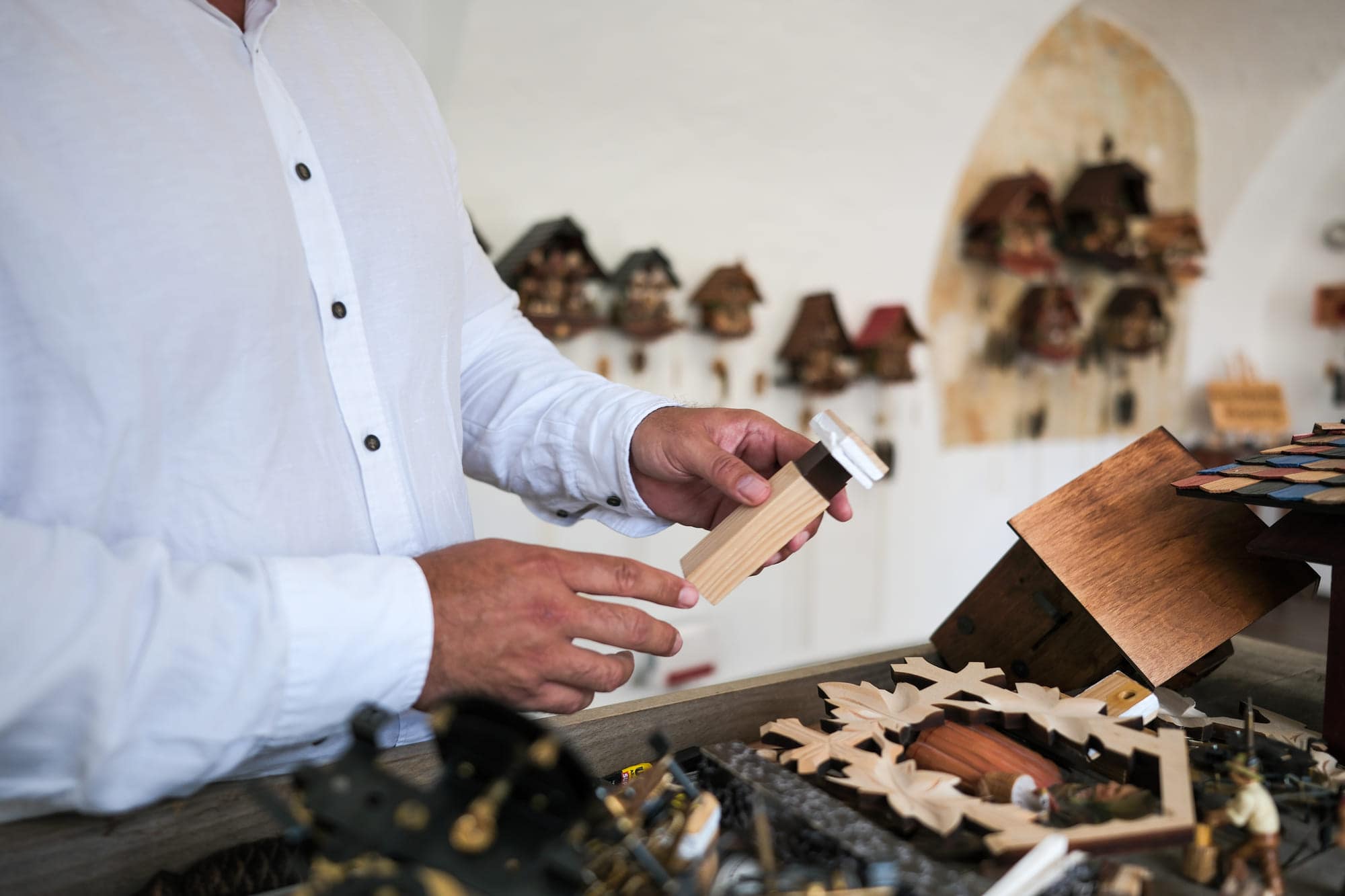 Zwei Hände halten einen rohen, unbehandelten Holzklotz, dahinter unscharf der Gewölbebogen und die Uhrenwand der InnResidenz in Passau.